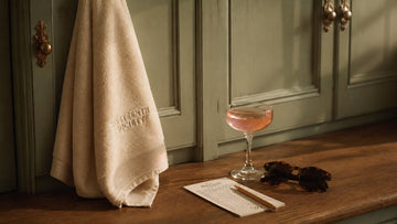 A locker room scene. A cream Nineteenth Society embroidered towel on an antique brass hook, a coupe glass with a pink cocktail, tortoise sunglasses, and a scorecard with a pencil on a warm wood counter. Sage green panelled cabinets in the background.