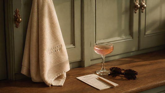 A locker room scene. A cream Nineteenth Society embroidered towel on an antique brass hook, a coupe glass with a pink cocktail, tortoise sunglasses, and a scorecard with a pencil on a warm wood counter. Sage green panelled cabinets in the background.